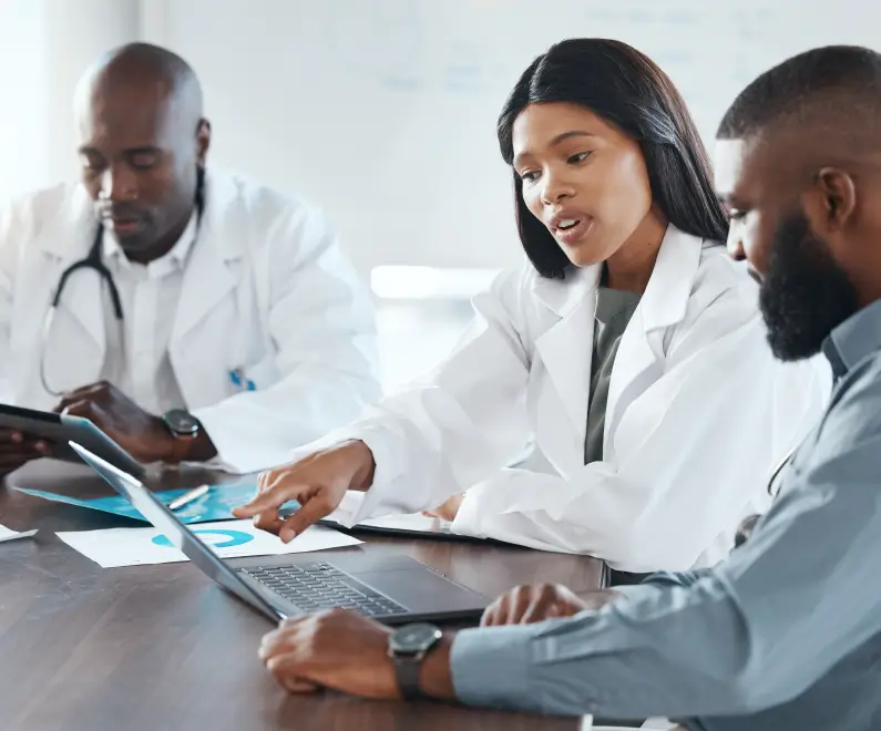 Group of three medical professionals sitting at table collaborating with paperwork and a laptop