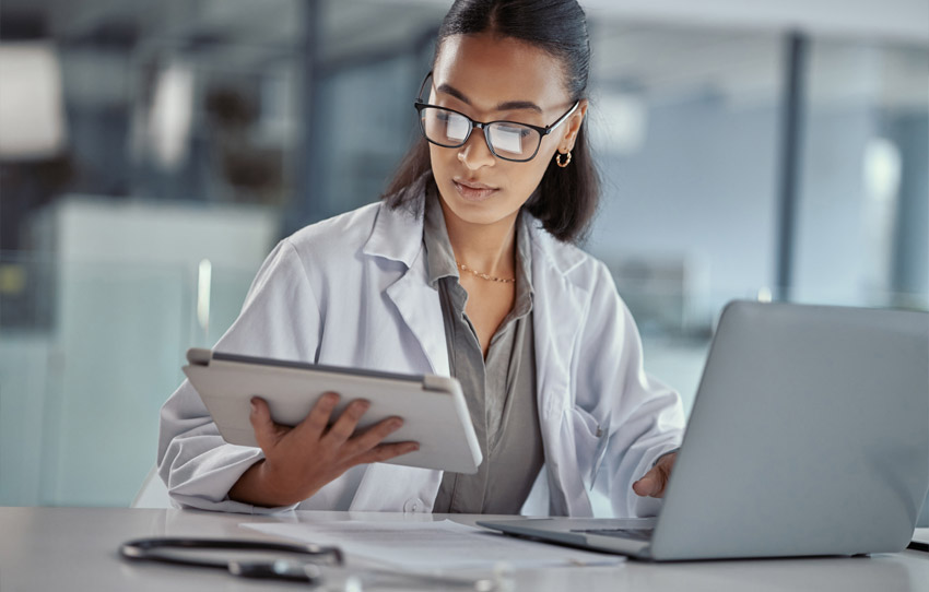 Medical professional reviews tablet at desk with laptop