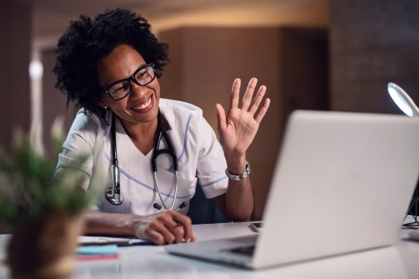 Black female healthcare worker waving and smiling while having video chat on the computer at doctor's office.