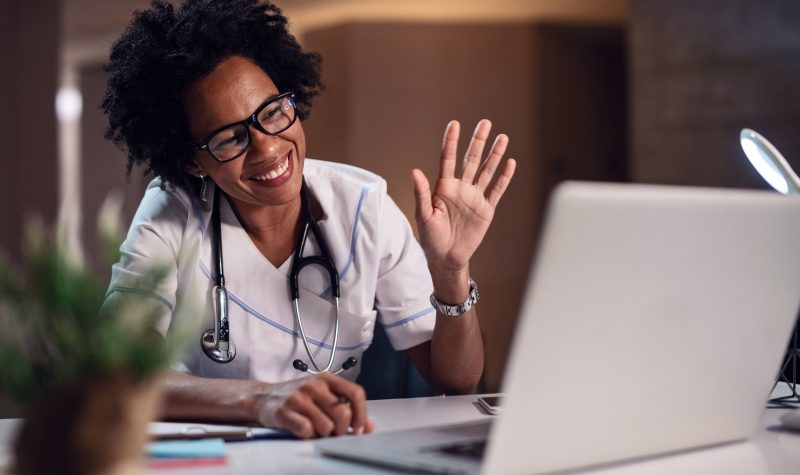 Black female healthcare worker waving and smiling while having video chat on the computer at doctor's office.