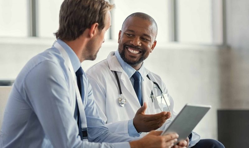 Two mature smiling doctors having discussion about patient diagnosis, holding digital tablet. Successful african surgeon discussing case after positive result. Representative pharmaceutical discussing with african happy doctor about new medicine.